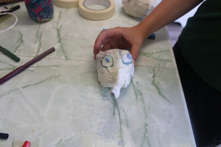 The head of a puppet with a big nose being currently made by one of the young people